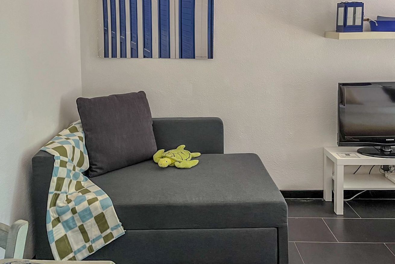 Ett modernt vardagsrum med en grå soffa, en grön, mjuk gosedjur, en rutig pläd, en platt-TV och ett vitt sidobord.