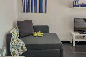 A modern living room with a grey sofa, a green plush toy, a checkered throw, a flat-screen TV, and a white side table.