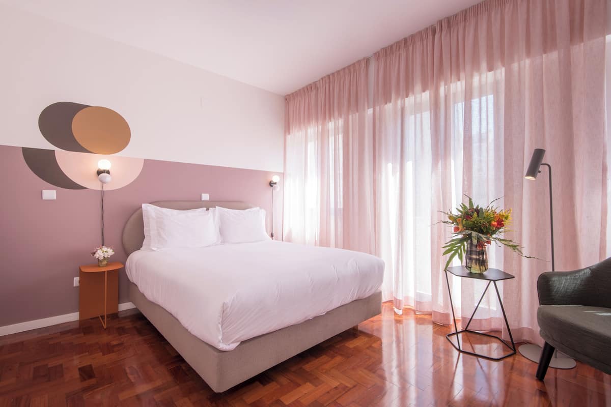 Ein modernes Schlafzimmer mit einem großen Bett, einem Nachttisch mit Lampe, einem kleinen Beistelltisch mit Blumenvase und einem grünen Sessel.