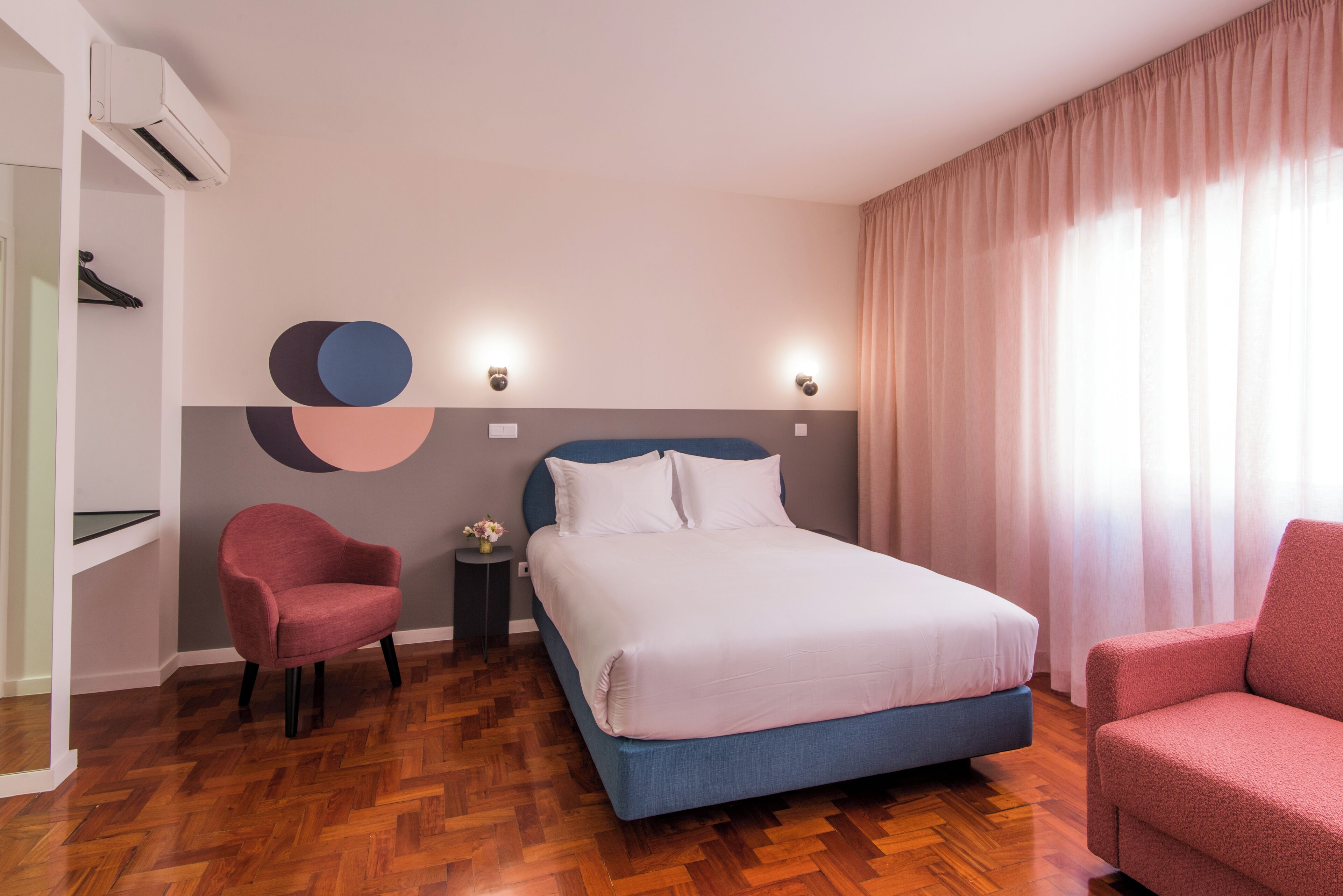 A hotel room with a bed, two chairs, a small table with a vase, and a wall-mounted air conditioner.