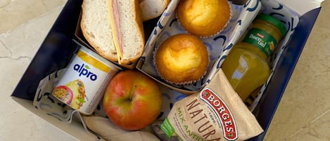 A lunch box containing sandwiches, muffins, a bottle of juice, a jar of peanut butter, a carton of almond milk, and an apple.