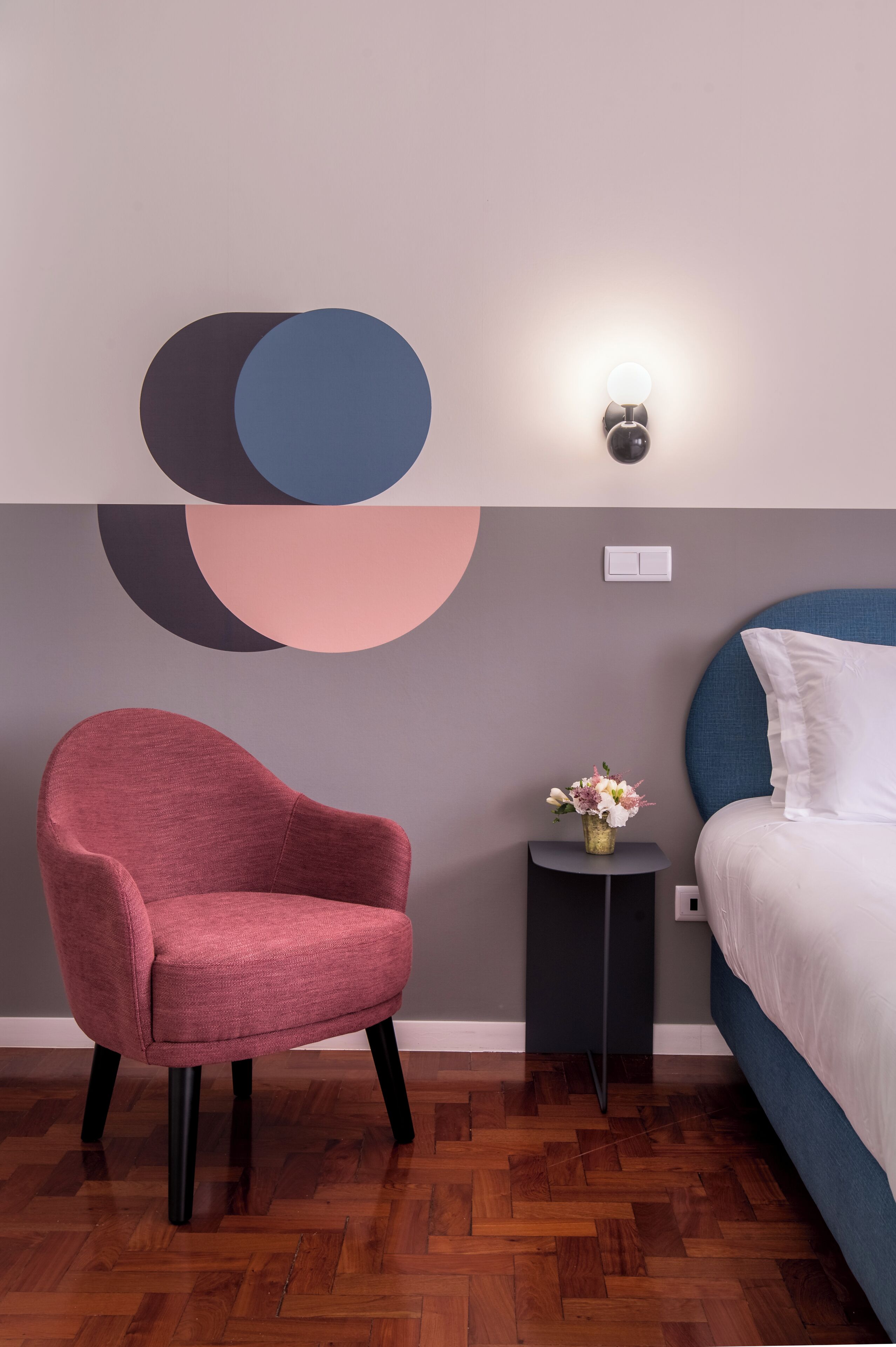 Une chambre moderne avec un parquet en bois disposé en motif chevrons, une chaise rouge ronde, une petite table ronde ornée d’un vase de fleurs et un lit avec une tête de lit bleue.