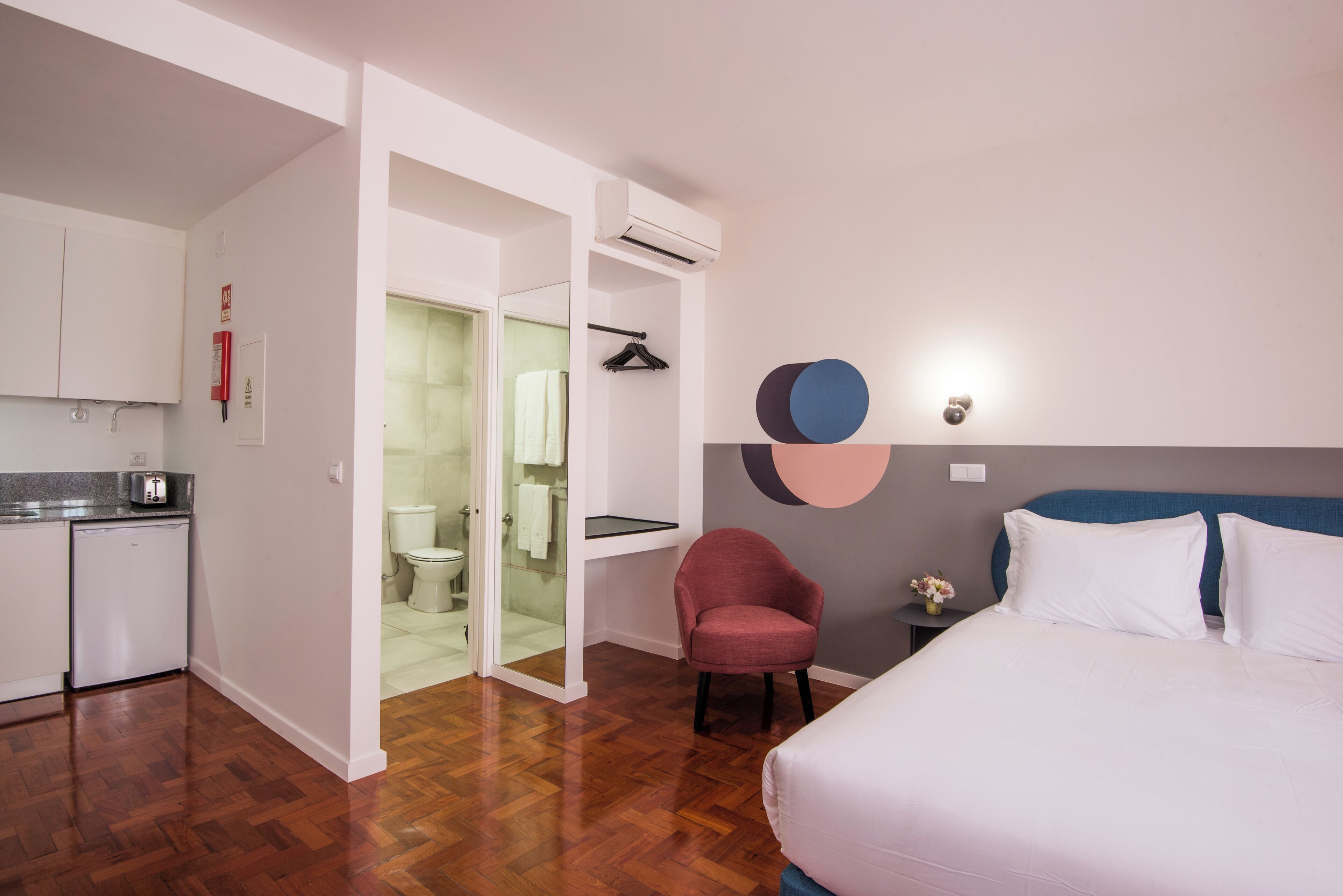 Une chambre d’hôtel compacte dotée d’une kitchenette, d’une salle de bain séparée avec une porte en verre, d’un lit avec du linge de lit blanc et d’une petite table de chevet agrémentée d’un vase.