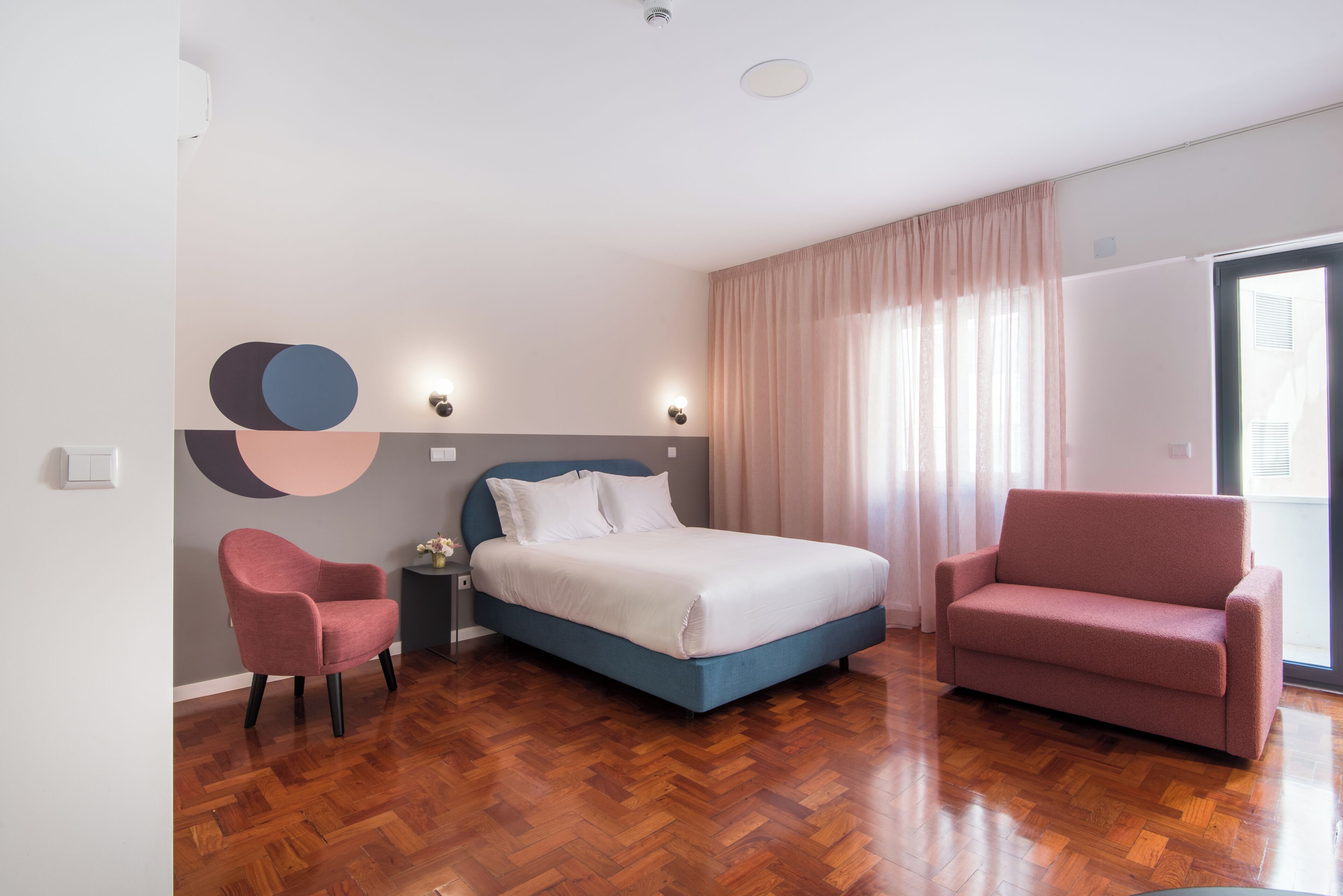 Une chambre à coucher moderne avec un lit, deux fauteuils, une table de chevet et un mur orné d’œuvres d’art abstraites.