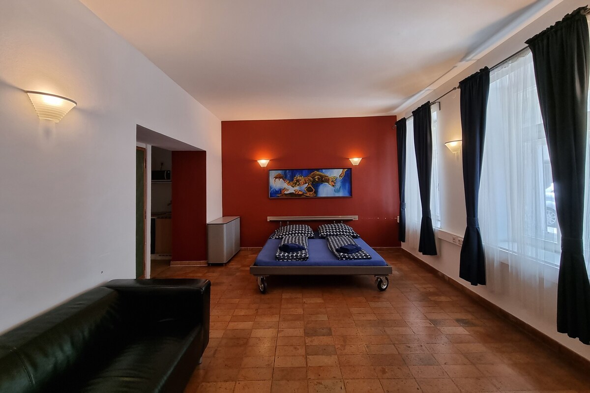 A room with a bed, a couch, a painting on the wall, and a window with curtains.