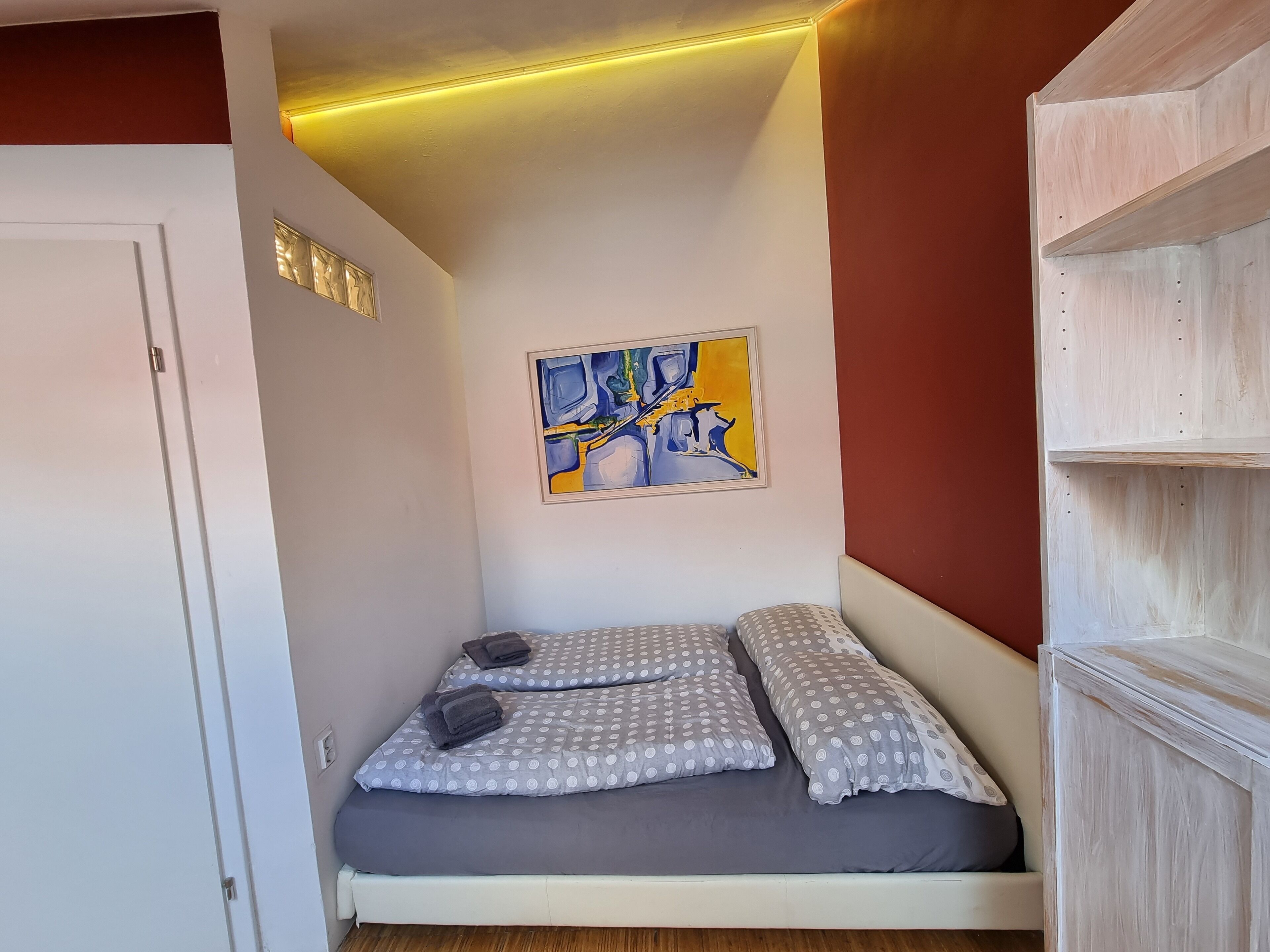 Una cama pequeña y bien hecha con ropa de cama gris y blanca a lunares, un cuadro abstracto colgado en la pared y un mueble estante de madera.