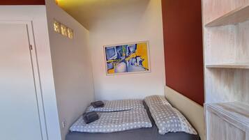 Una cama pequeña y bien hecha con ropa de cama gris y blanca a lunares, un cuadro abstracto colgado en la pared y un mueble estante de madera.
