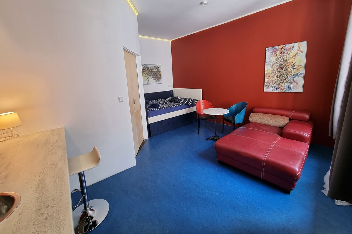 A room with a bed, a red sofa, a small table, and chairs. There is a painting on the wall and a lamp on the left.