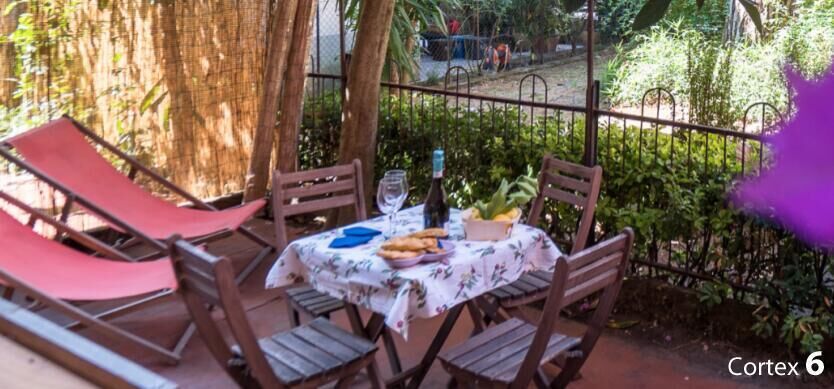 A patio with a table set for two, surrounded by wooden chairs, a red lounge chair, and a tablecloth.