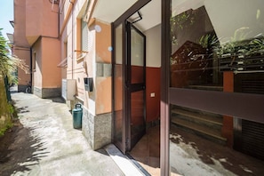 Une entrée d’immeuble avec une porte en verre, un chemin en béton et une poubelle verte.