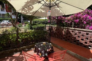 Un espace extérieur aménagé avec une table et des chaises, protégé par un grand parasol, et entouré de verdure et d’un mur de briques.