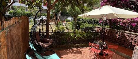 Un área de estar al aire libre con sillas y mesas, rodeada de vegetación y un edificio al fondo.
