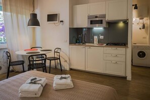 A compact hotel room with a kitchenette, a dining area with a table and chairs, a bed with towels, and a washing machine.