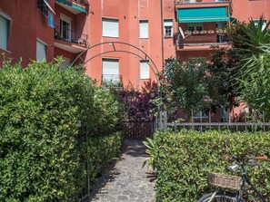 Un étroit sentier de pierre, bordé de haies verdoyantes et touffues, mène à un bâtiment doté de balcons et d’un mur en briques.