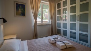 Une chambre à coucher avec un lit, une fenêtre avec des rideaux, une lampe fixée au mur et une porte avec des panneaux de verre dépoli.