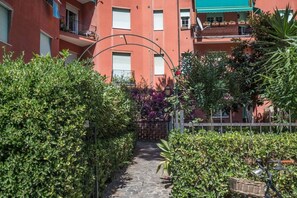 A narrow stone pathway flanked by dense green hedges and a red building with balconies.