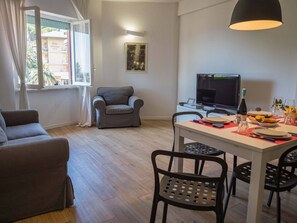 Un salon comprenant un canapé, une table à manger avec des chaises, un téléviseur et une fenêtre avec des rideaux.