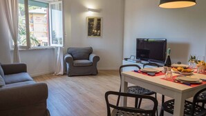 A living room with a sofa, a grey armchair, a dining table with chairs, a flat-screen TV, and a window with curtains. - Scimiscià 2 bedroom Apartment (Monterosso al Mare)
