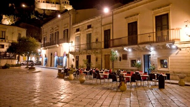 Un área de café al aire libre con mesas y sillas, un edificio con balcón, y un edificio histórico al fondo.