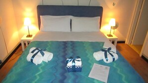 Habitación de hotel con cama, dos lámparas de noche, una pared decorada con motivos florales y una colcha con una cesta de regalo y una nota.