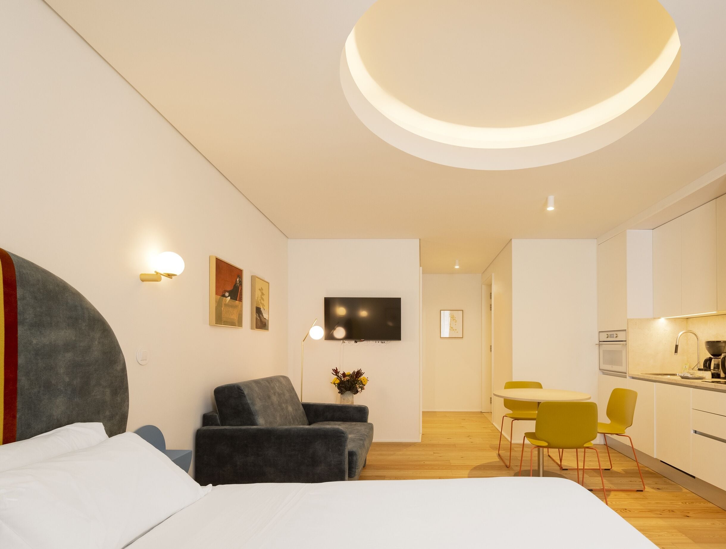 A modern hotel room with a bed, a sofa, a dining area with chairs, a TV, and a kitchen area.