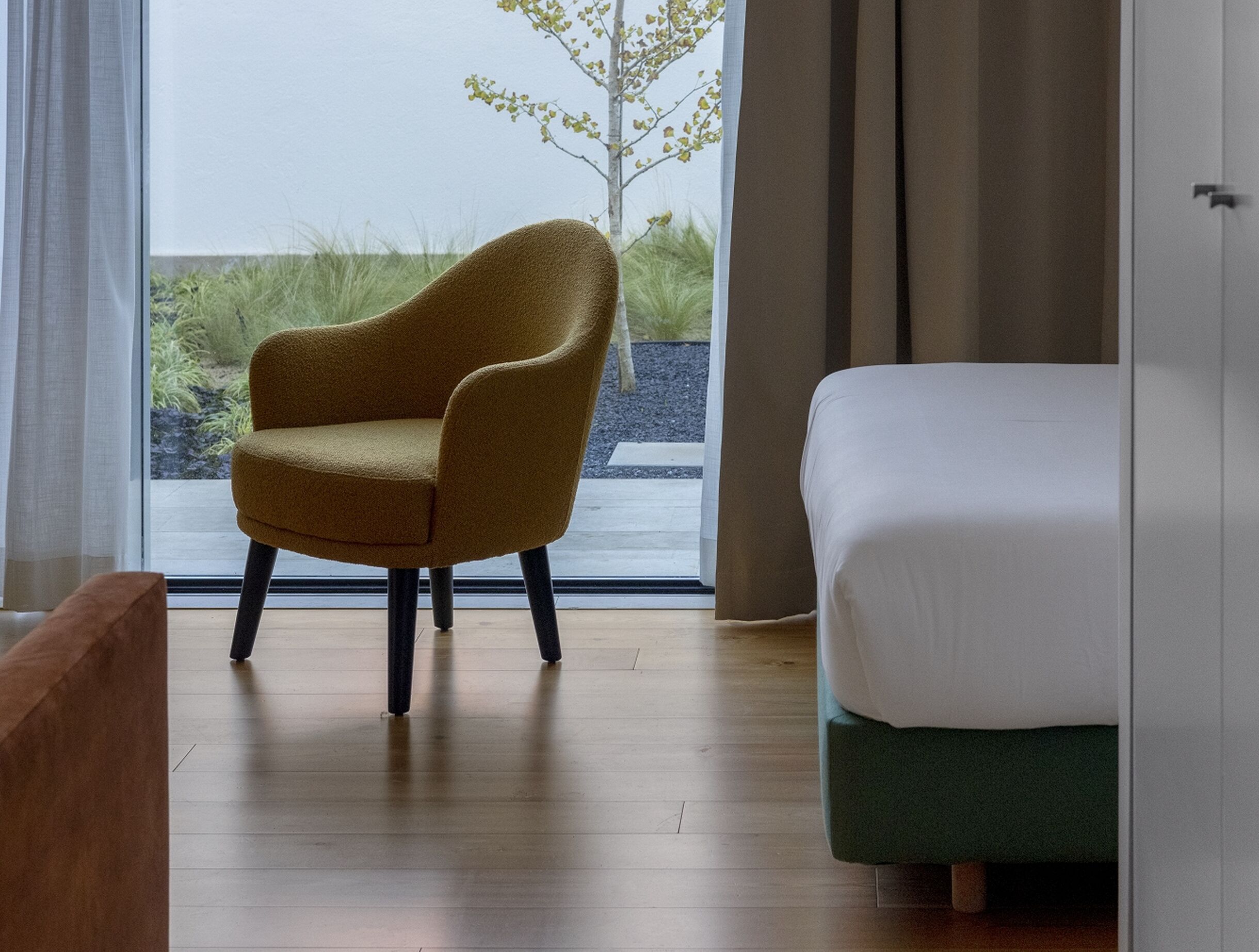 A room with a bed, a chair, and a window with a view of grass and a tree.