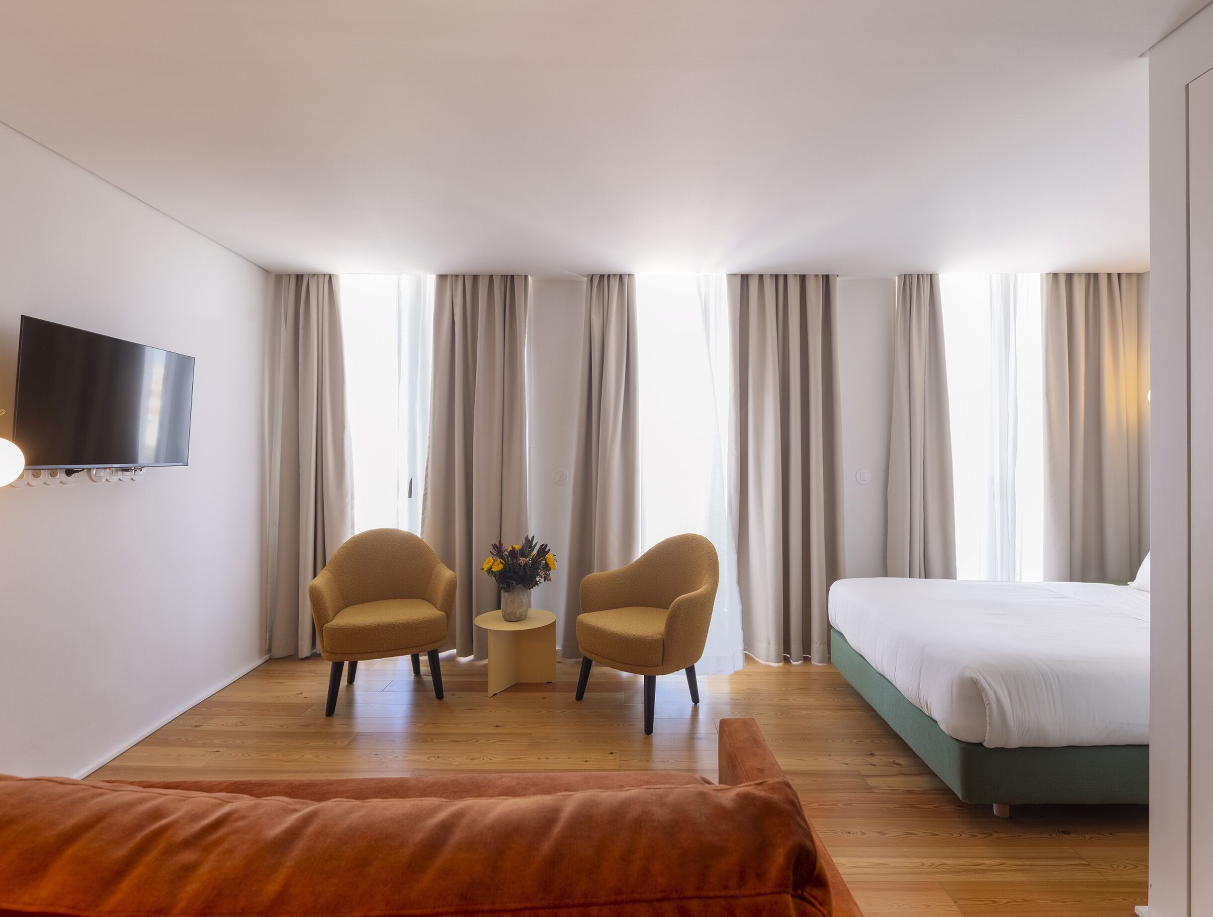 Una camera d'albergo con un letto, due poltrone, un tavolino con un vaso, una televisione a schermo piatto montata a parete e ampie finestre con tende.