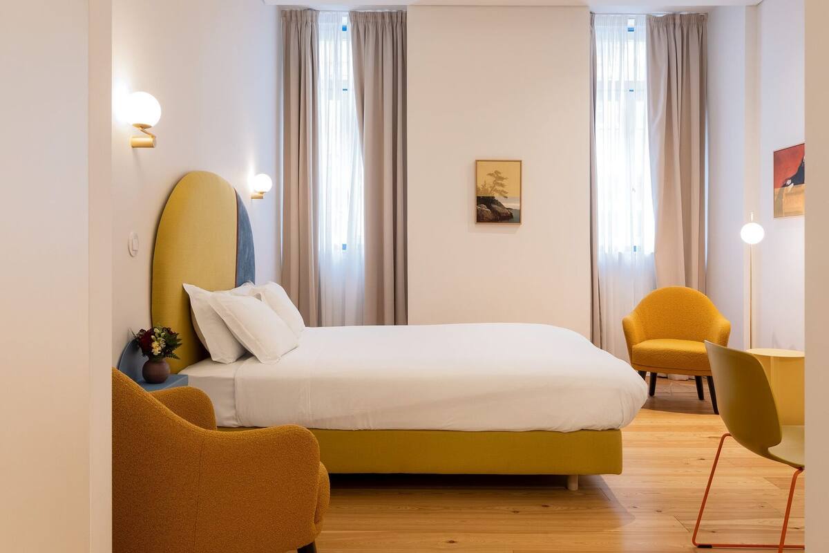 A hotel room with a bed, two chairs, a desk, and a painting on the wall.