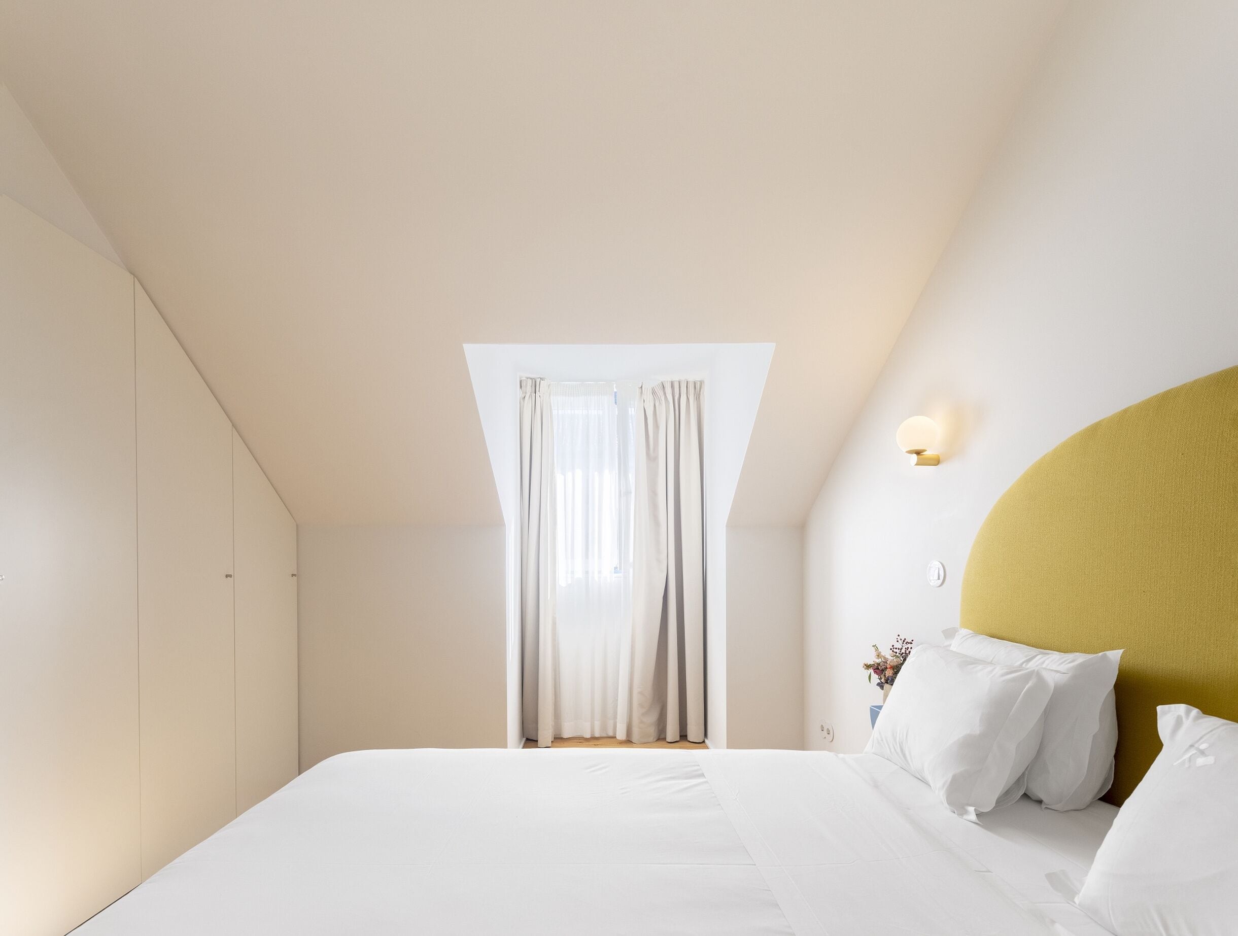 Een slaapkamer met een bed, een raam met gordijnen, een wandlamp en een gele accentmuur.