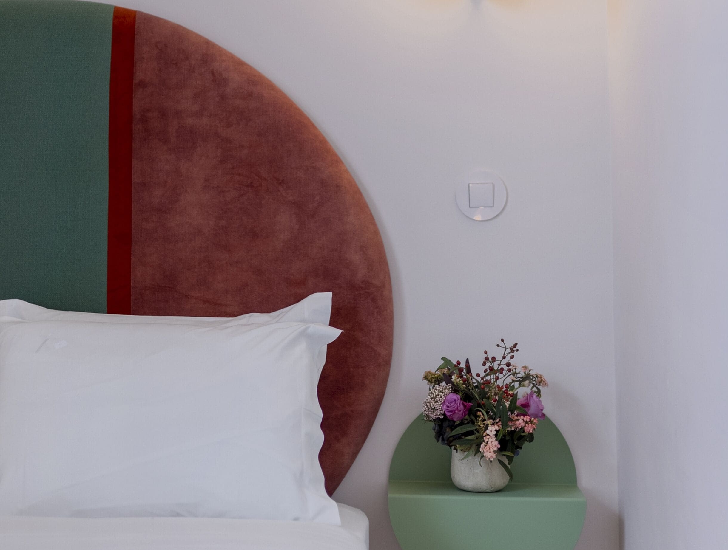 Een bed met witte beddengoed, een rond hoofdbord met een rode streep, een nachtkastje in het groen met een bloevas, en een lichtschakelaar aan de muur.