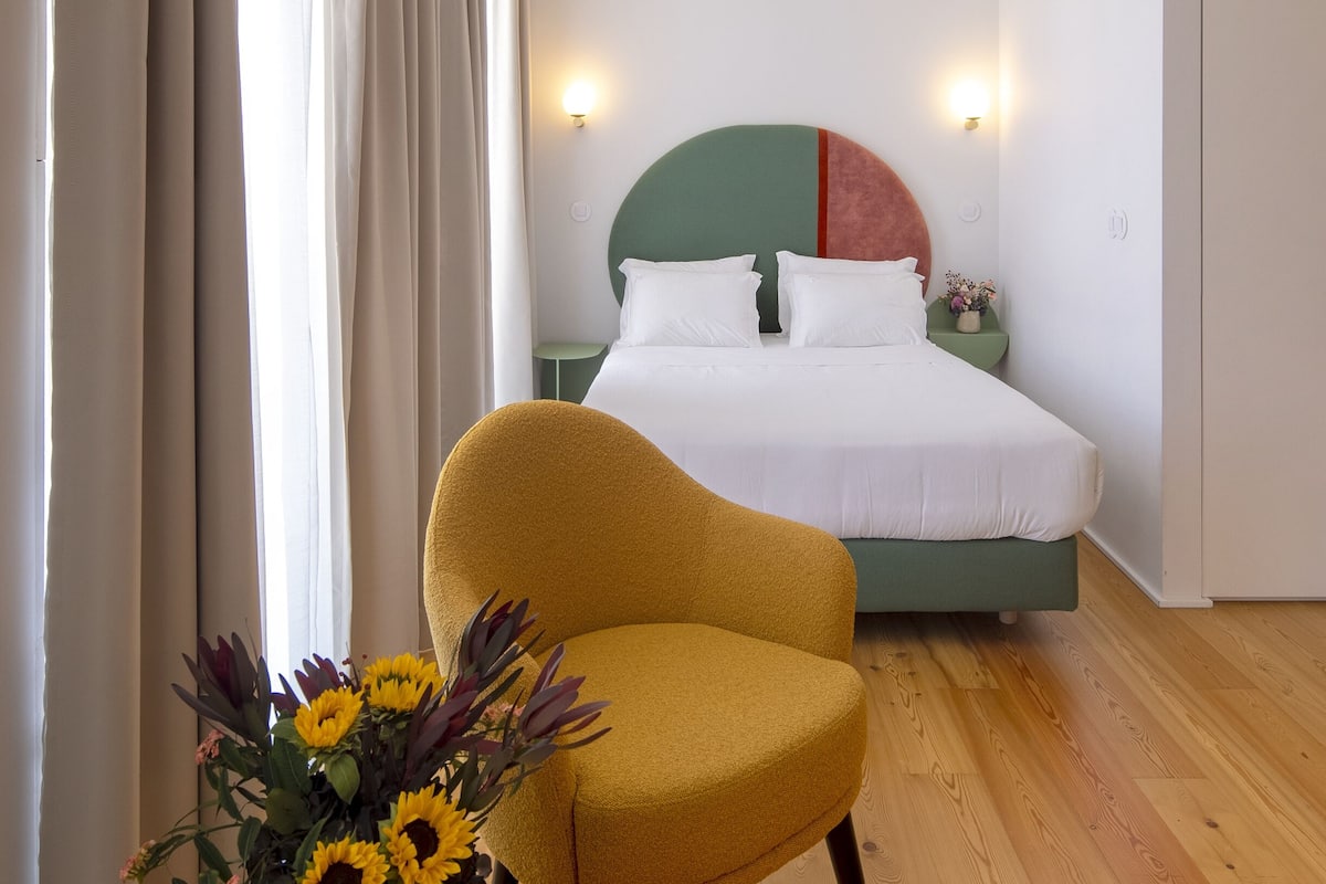 A hotel room with a bed, a yellow armchair, a nightstand, and a wall-mounted light.