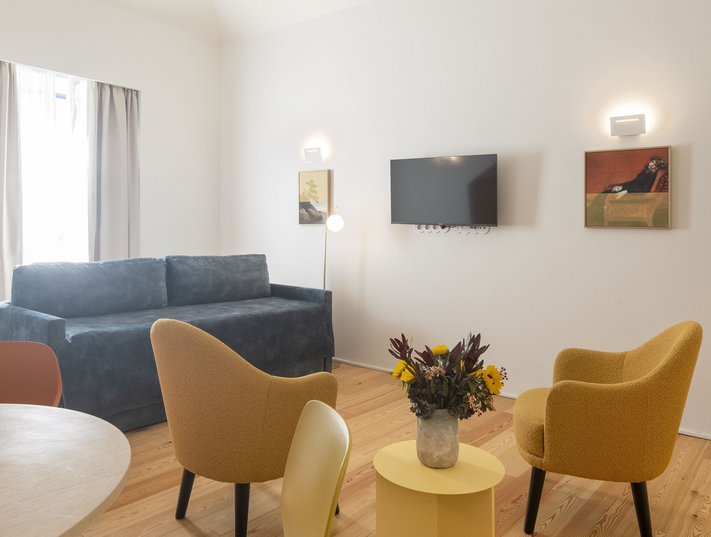 A modern living room with a grey sofa, two mustard-colored chairs, a small yellow table with a vase of flowers, a flat-screen TV mounted on the wall, and a painting.