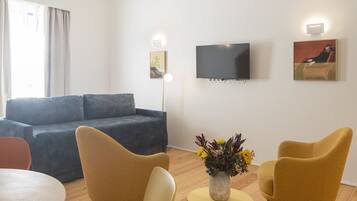 A modern living room with a grey sofa, two mustard-colored chairs, a small yellow table with a vase of flowers, a flat-screen TV mounted on the wall, and a painting.