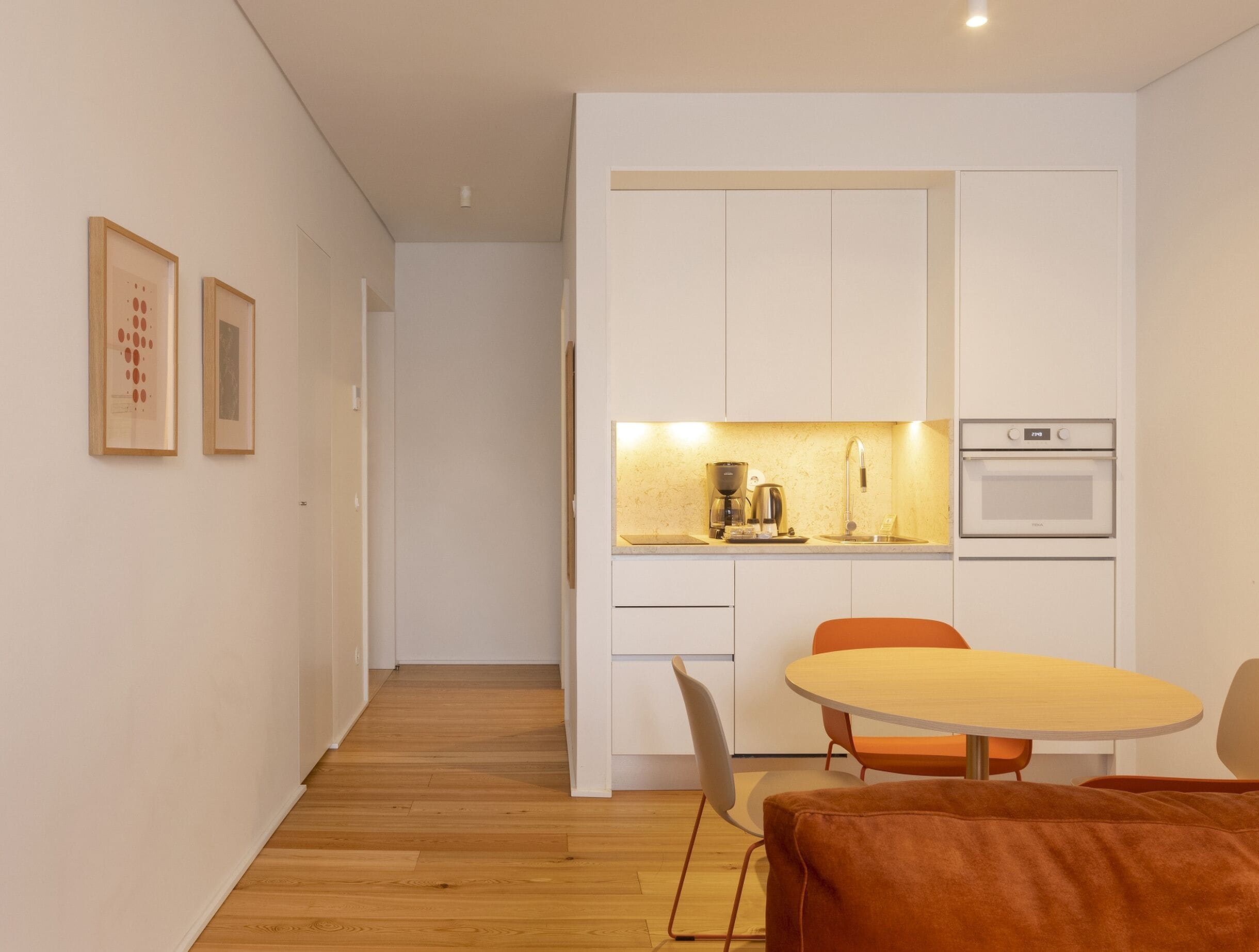 Una cocina moderna con armarios blancos, una cafetera y un horno. Hay una zona de comedor con una mesa redonda y sillas naranjas.