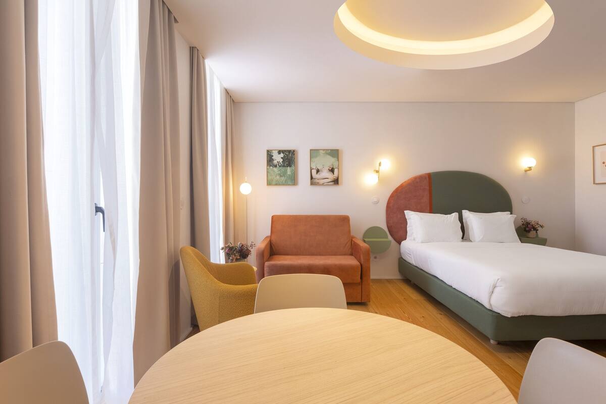 A hotel room with a bed, a round table, a yellow chair, a brown armchair, and a green headboard.
