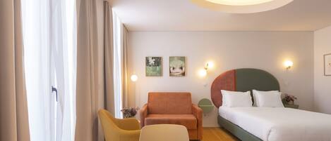 A hotel room with a bed, a round table, a yellow chair, a brown armchair, and a green headboard.
