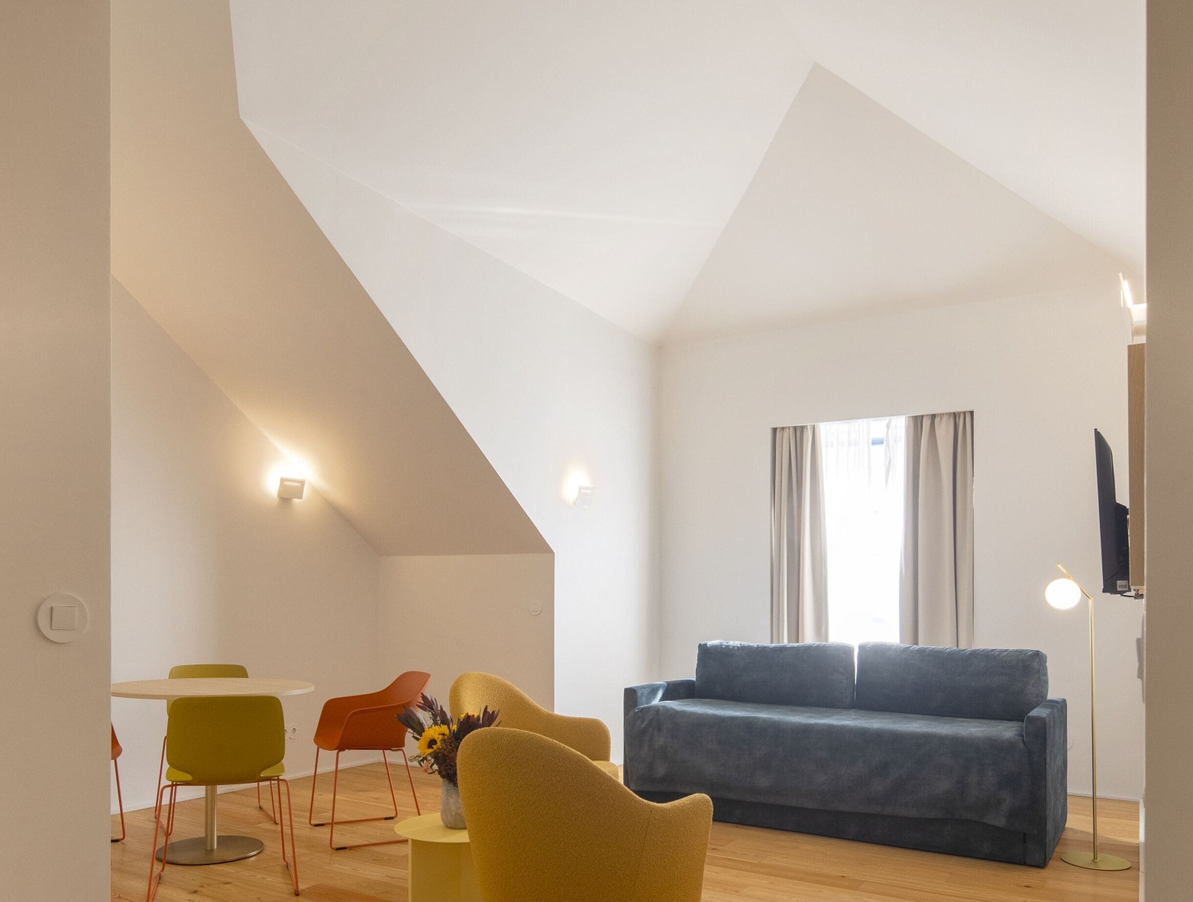 Un soggiorno moderno con soffitto alto, un divano blu, poltrone gialle e arancioni, un tavolino con un vaso e una TV a parete.