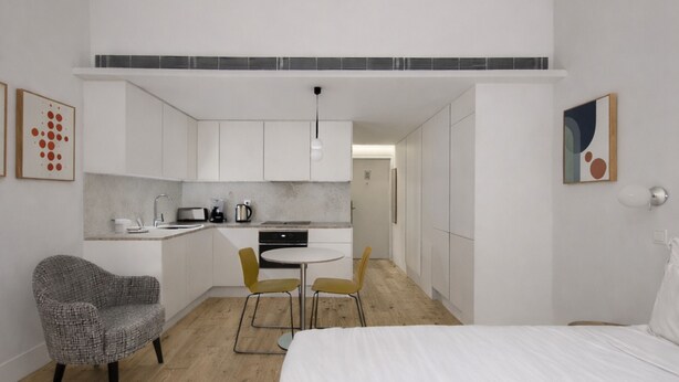 A compact hotel room with a bed, a small kitchenette, a dining area with chairs, and a gray patterned armchair.