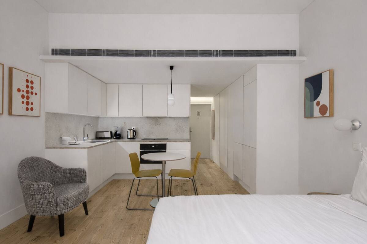 Habitación de hotel compacta con cama, pequeño rincón de cocina, comedor con sillas y un sillón gris estampado.