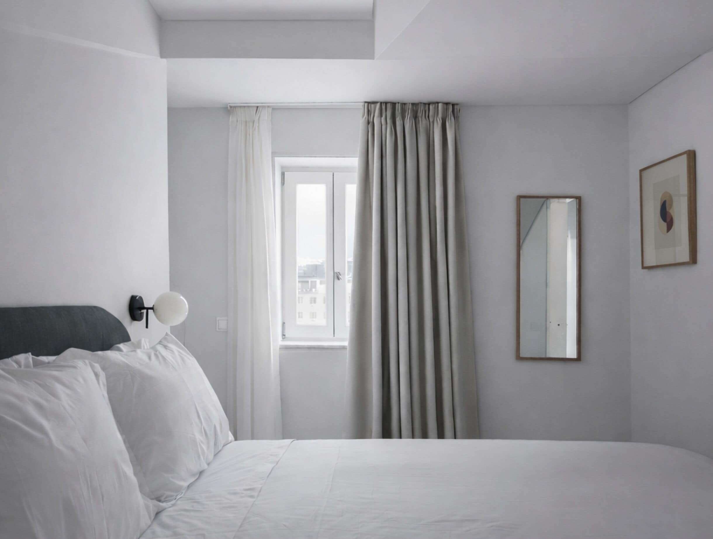 A minimalist hotel room with a bed, a window with curtains, a wall-mounted mirror, and a framed picture.