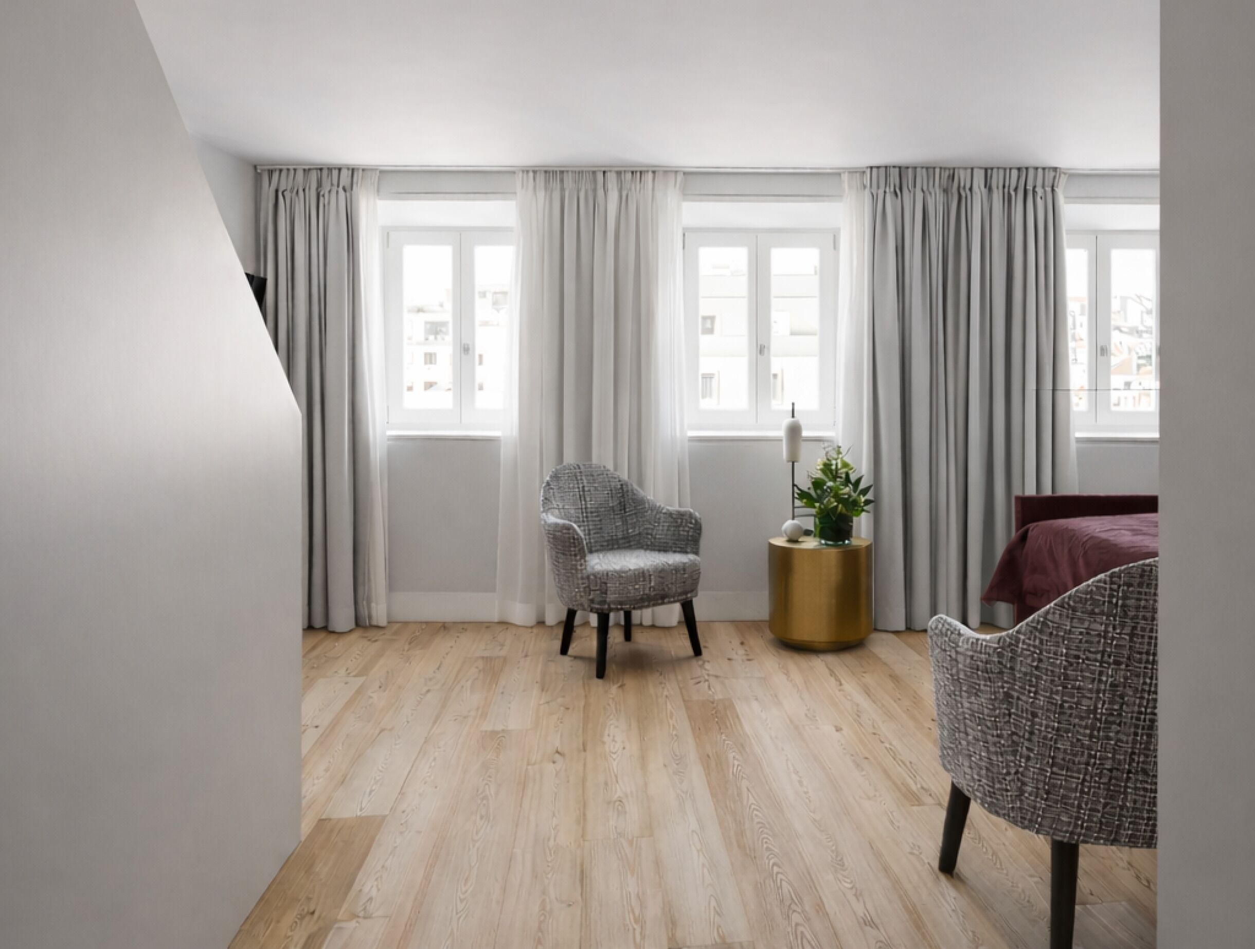 Een moderne slaapkamer met een houten vloer, een grijze fauteuil met een patroon, een rond goudkleurig bijzettafeltje met een plant, en een bed met een rode spreidekker.