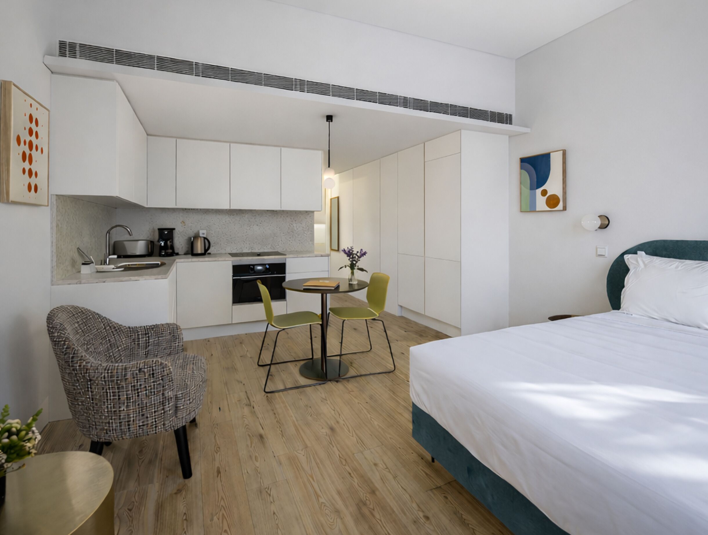 A modern hotel room with a bed, a dining table with chairs, a kitchenette, and a small seating area.