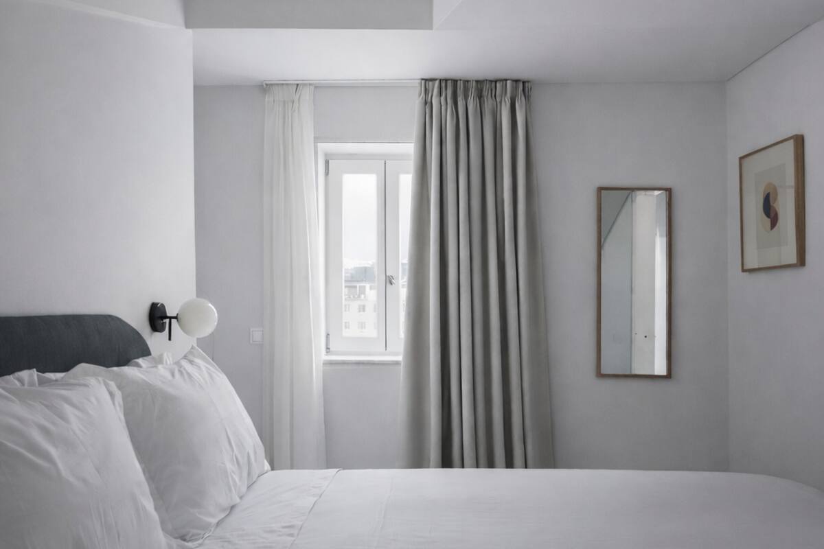 Habitación de hotel minimalista con una cama, una lámpara de noche, una ventana con cortinas y un espejo en la pared.