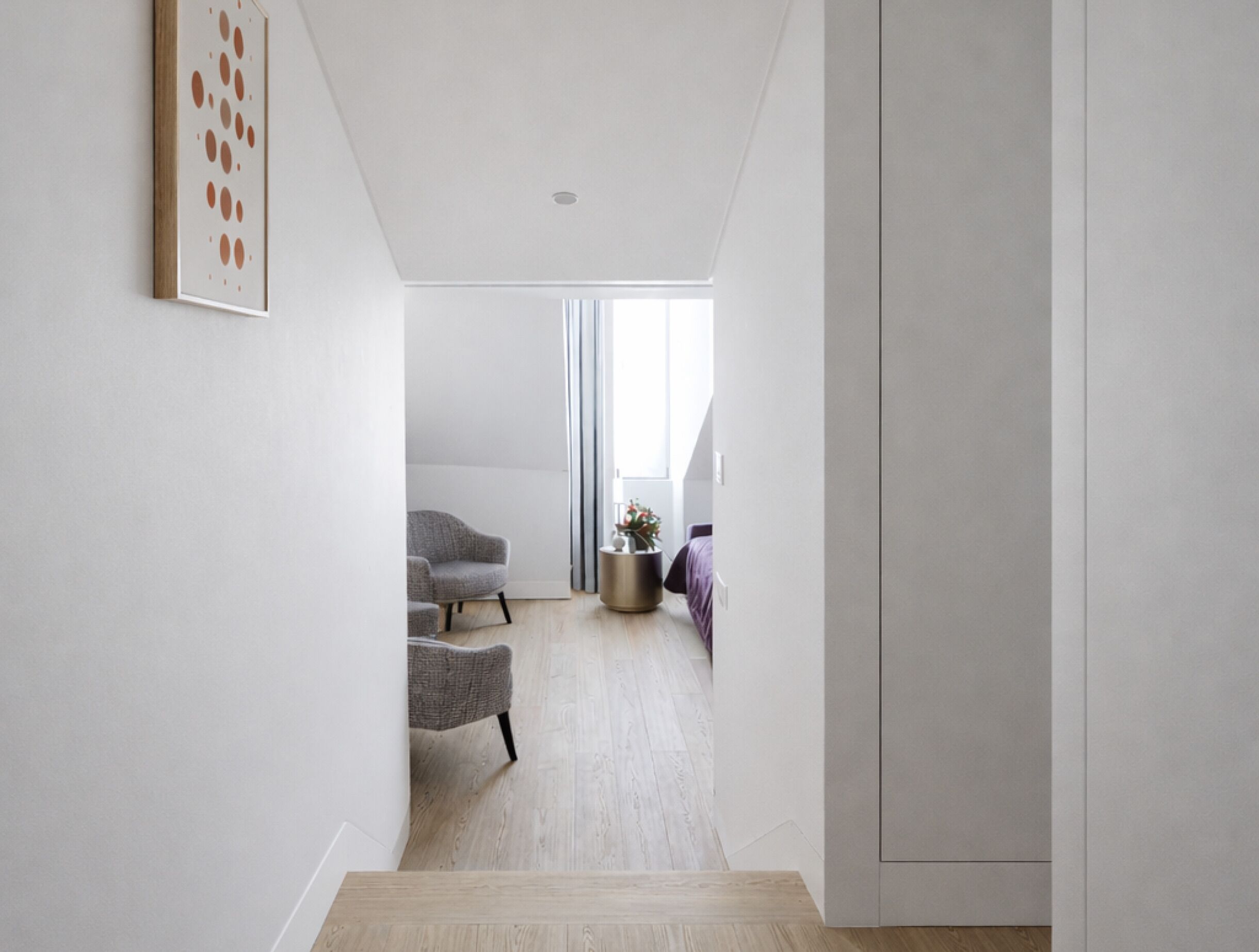 Un couloir étroit avec un escalier menant à une pièce dotée d’une fenêtre, d’une chaise et d’une petite table sur laquelle se trouve une plante.