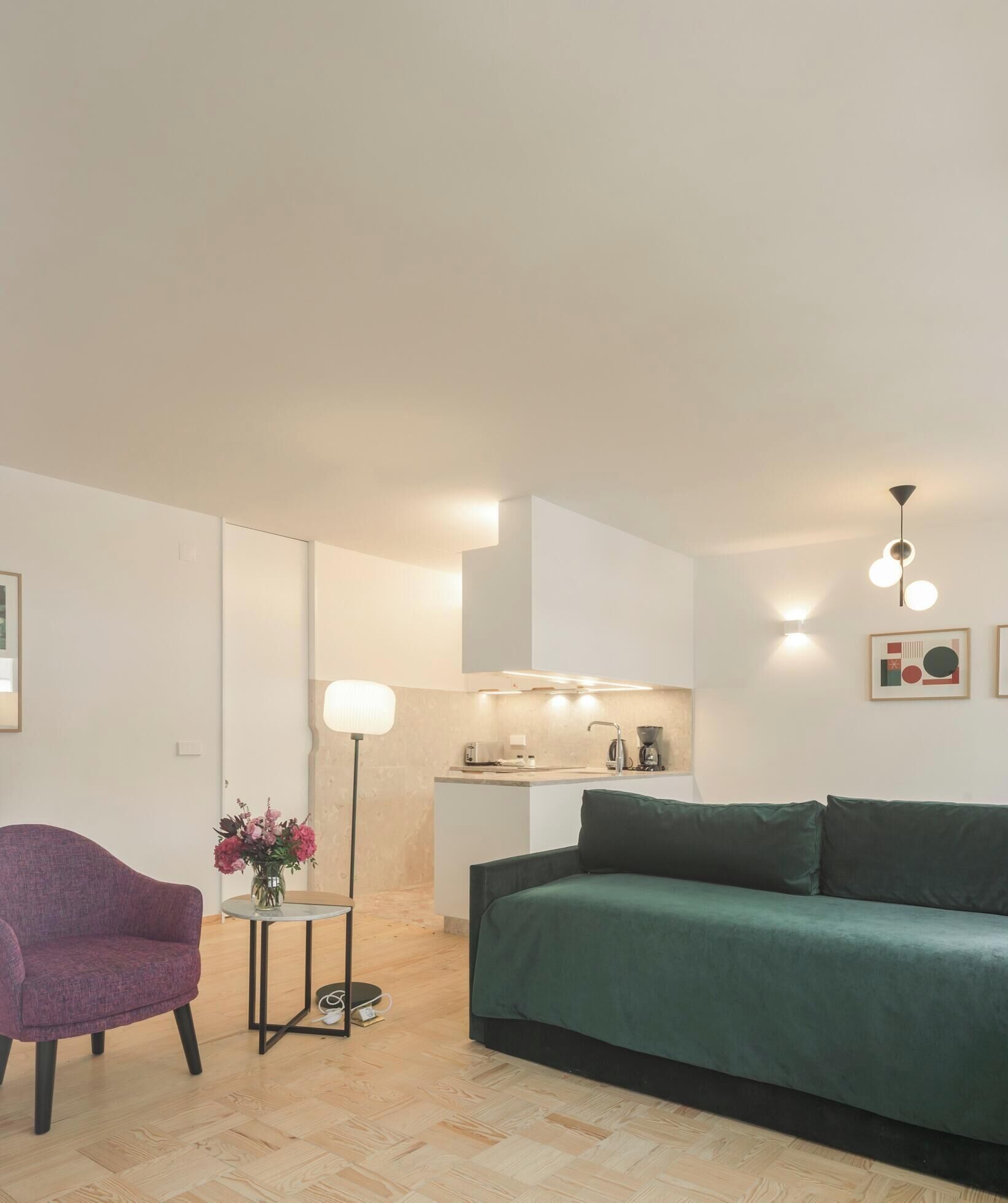 Een moderne woonkamer met een groene bank, een paarse fauteuil, een bijzettafeltje met een bloevas, en een keuken in de achtergrond.
