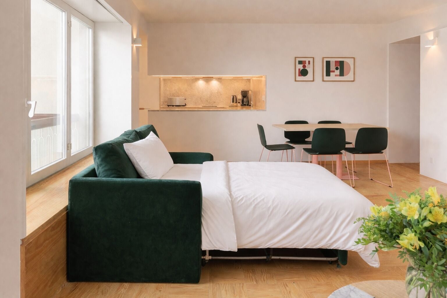 A modern room with a green sofa, a bed, a dining table with chairs, and a small kitchenette.