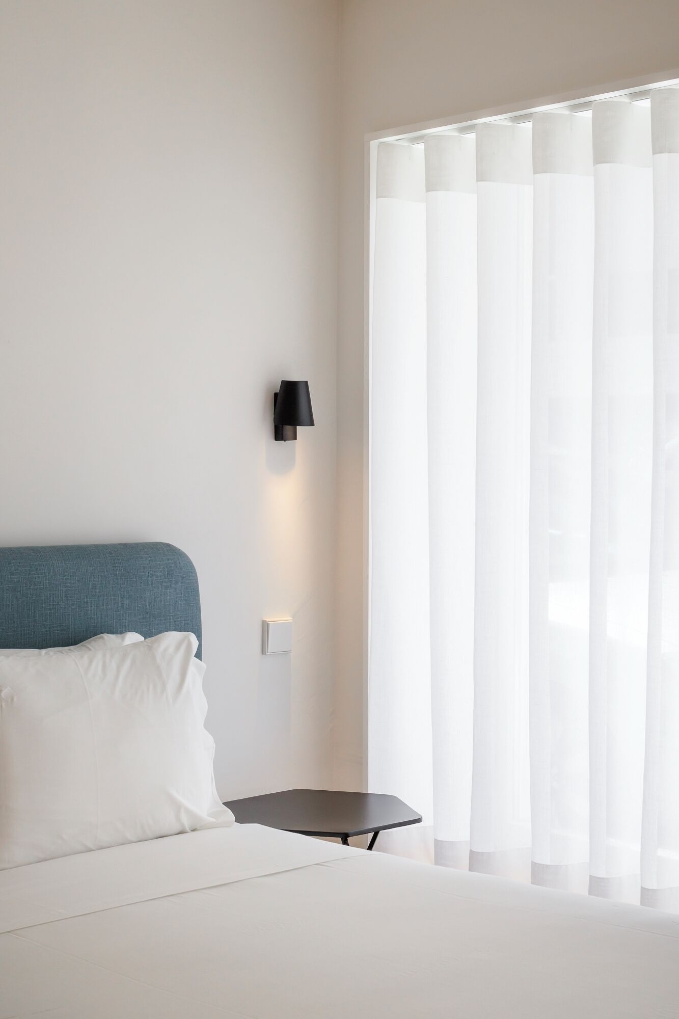 A modern hotel room with a bed, a small table, a wall-mounted lamp, and sheer curtains.