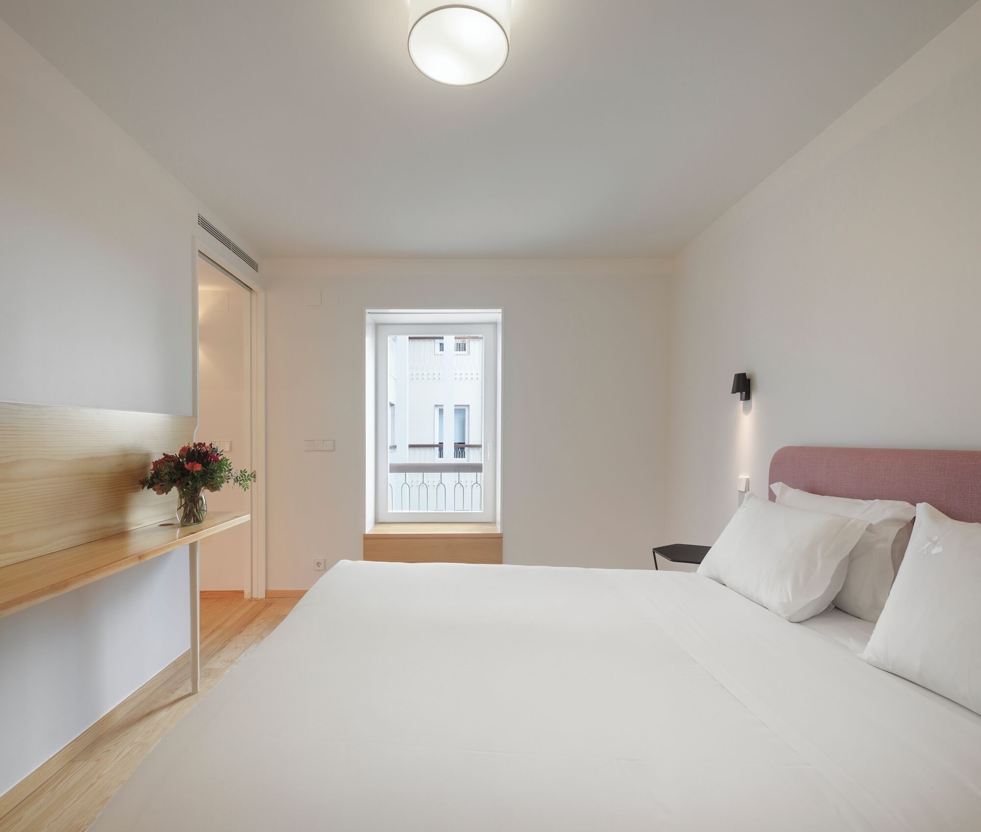 Un dormitorio con cama, una mesita de noche, una lámpara de pared, un ventanal con vistas y un estante de madera con flores.