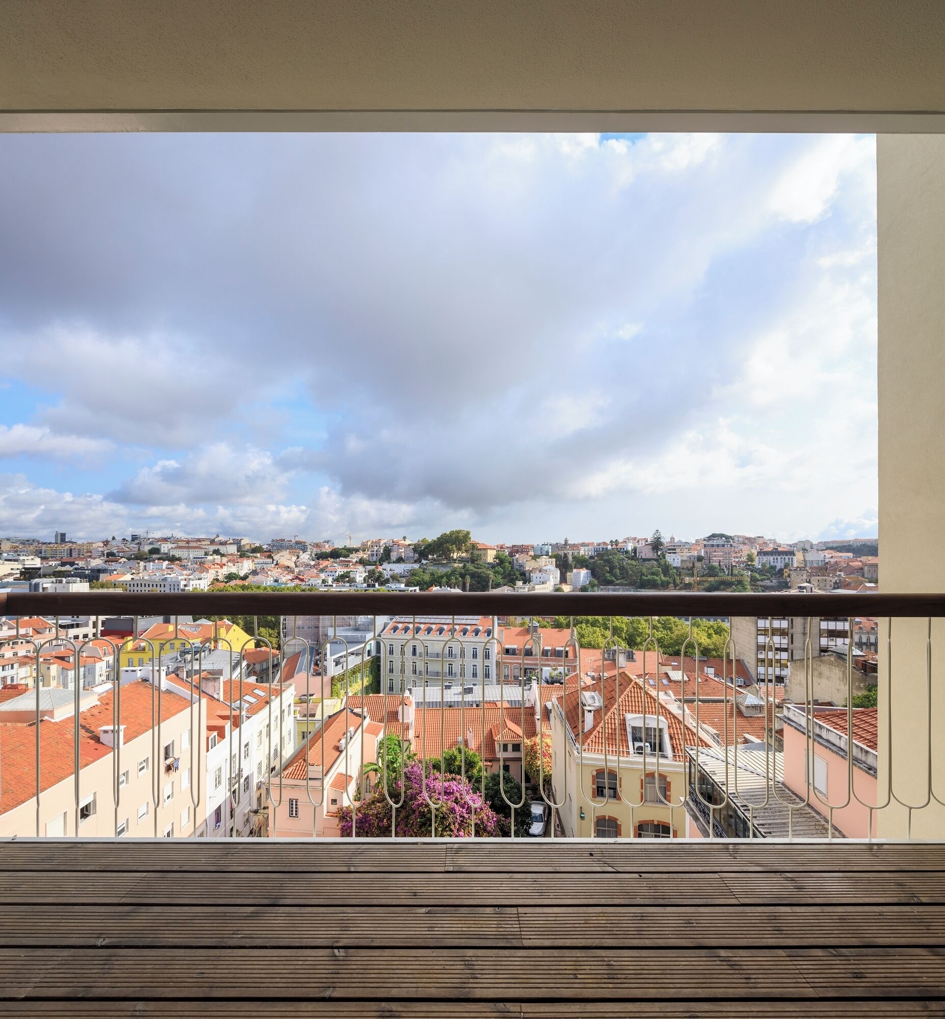 Un balcon offrant une vue sur le paysage urbain, avec des bâtiments aux toits recouverts de tuiles rouges et un ciel nuageux.