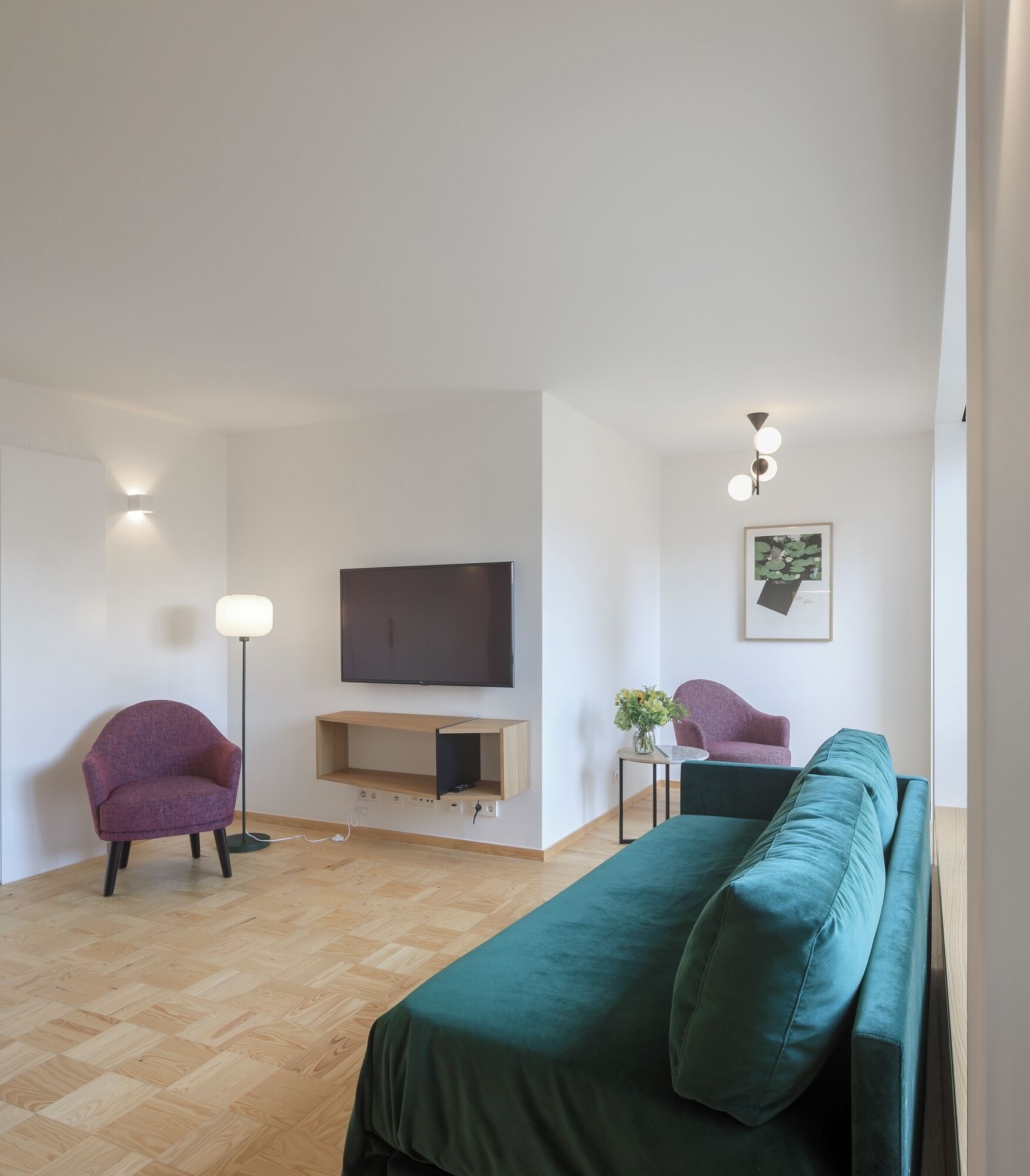 Un salon moderne avec un téléviseur à écran plat, un canapé vert, un fauteuil violet, une lampe sur pied et une petite table sur laquelle est posé un vase.